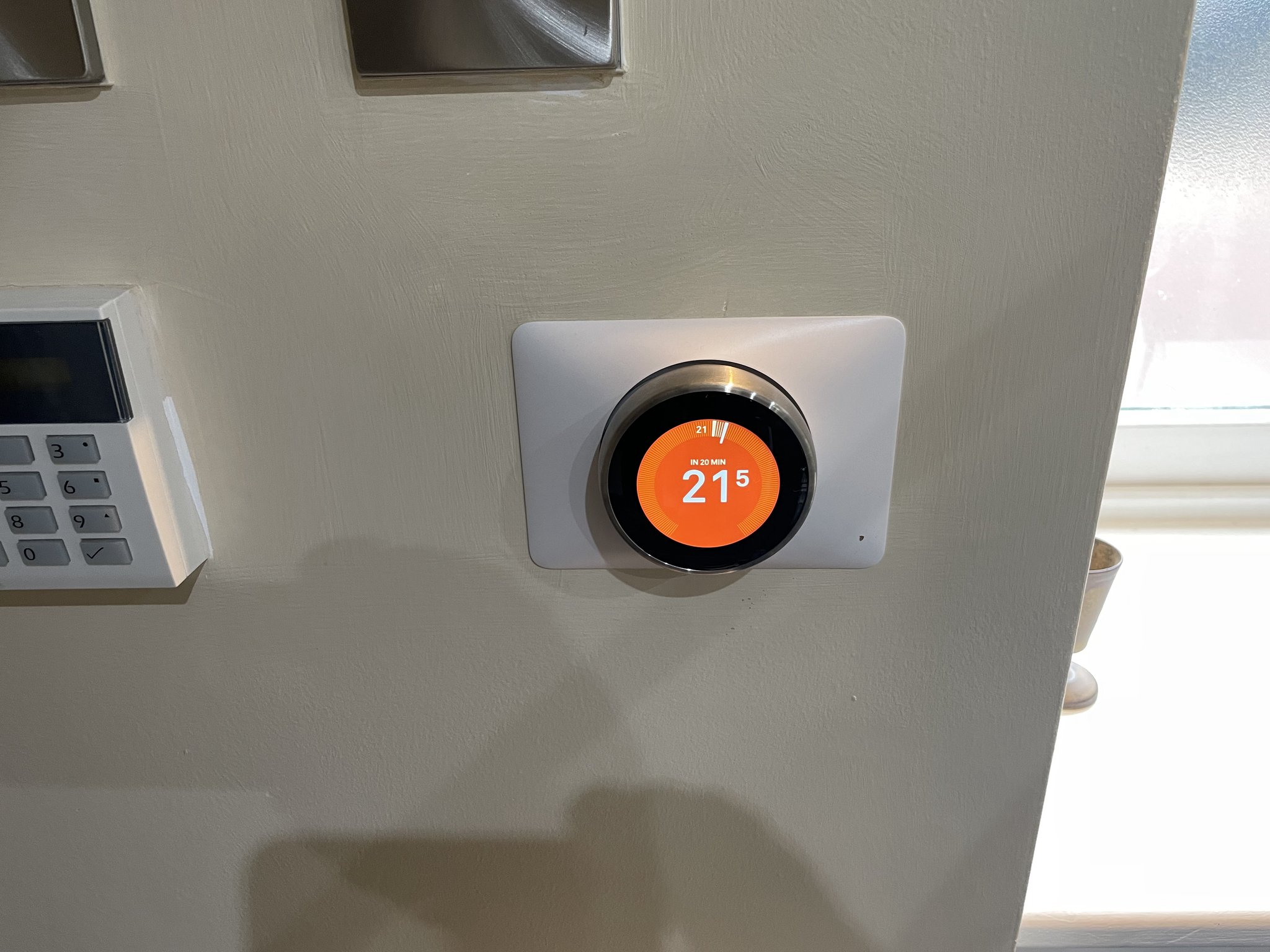 A Nest thermostat fitted on a grey wall beside an alarm keypad